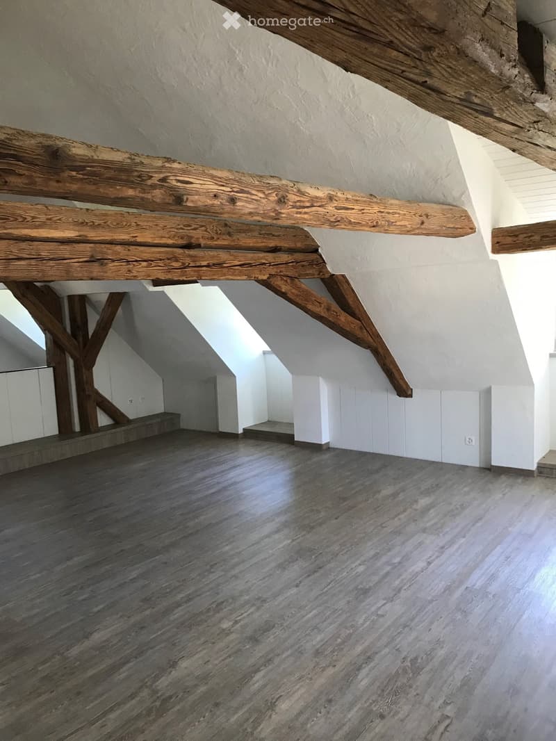Empty attic space with exposed wooden beams, wooden flooring, and white walls. No furniture or equipment present.
