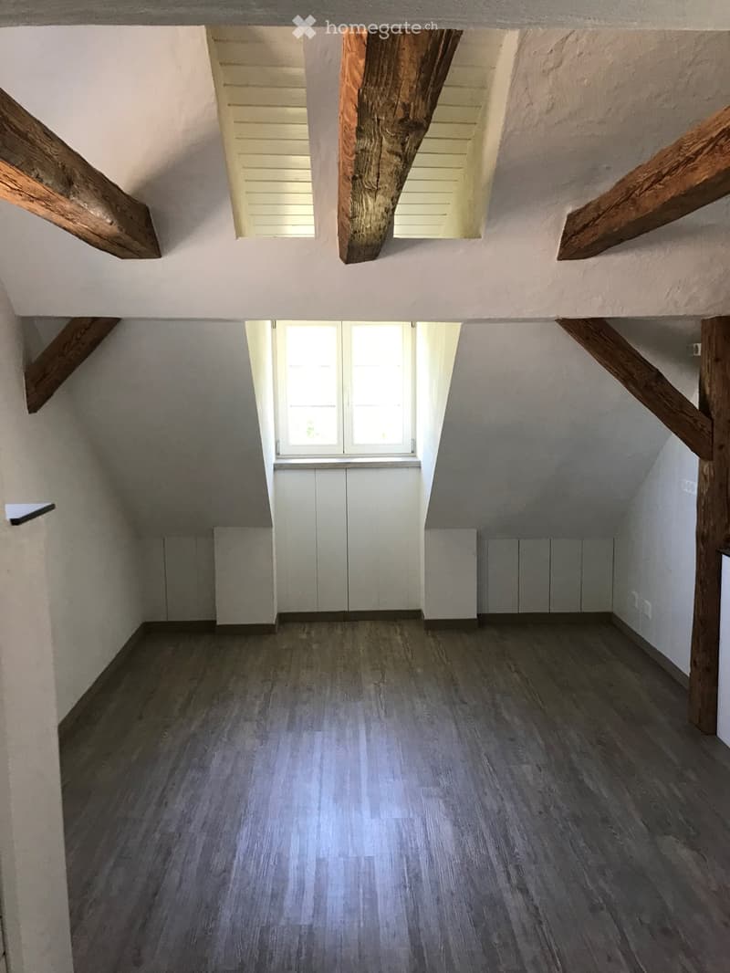 Empty attic space with wooden beams, exposed wooden ceiling, and a window letting in natural light. The floor is covered with light grey wooden planks.