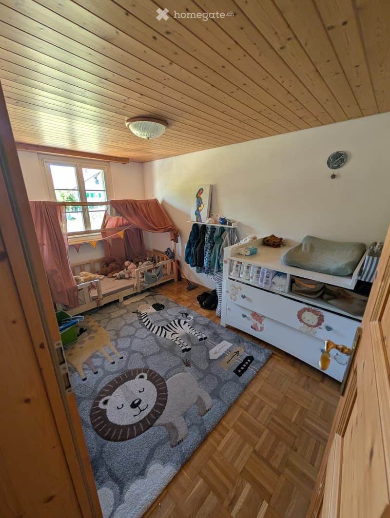 Cozy children's bedroom with wooden floor, slanted wooden ceiling, large window with curtain, bed with canopy, storage unit, and animal-themed rug.