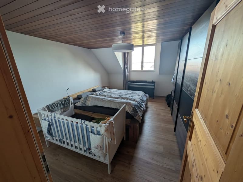 Cozy bedroom with wooden floors, slanted ceiling, bed, cradle, piano, and a hanging lamp.