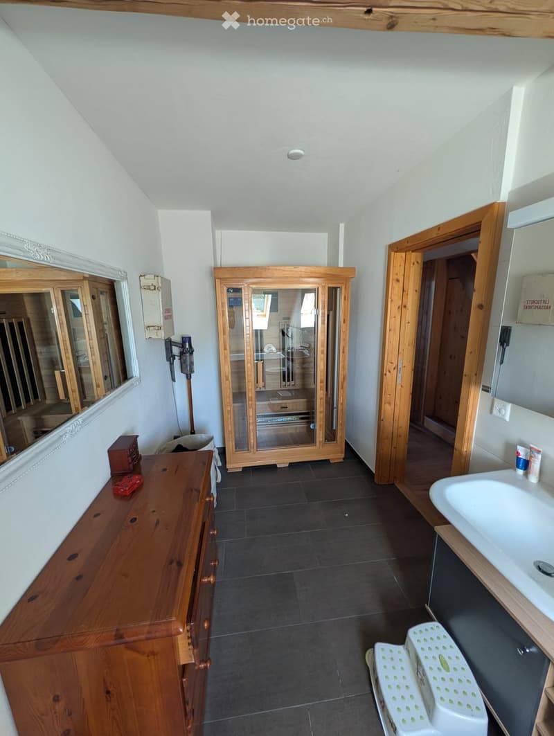 Bathroom with dark tiled floor, white walls, wooden cabinet, sink, mirror, and a small stool near the sink.