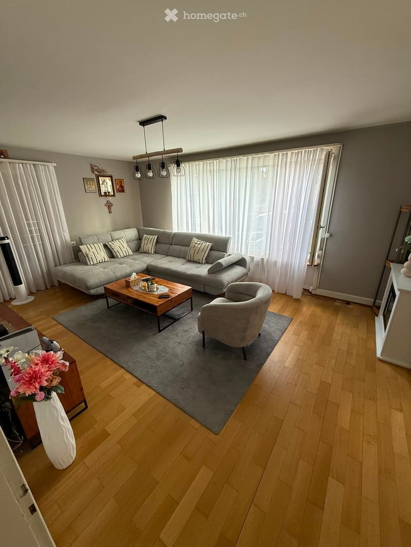 Modern living room with wooden flooring, grey sectional sofa, coffee table, armchair, and large window with white curtains.