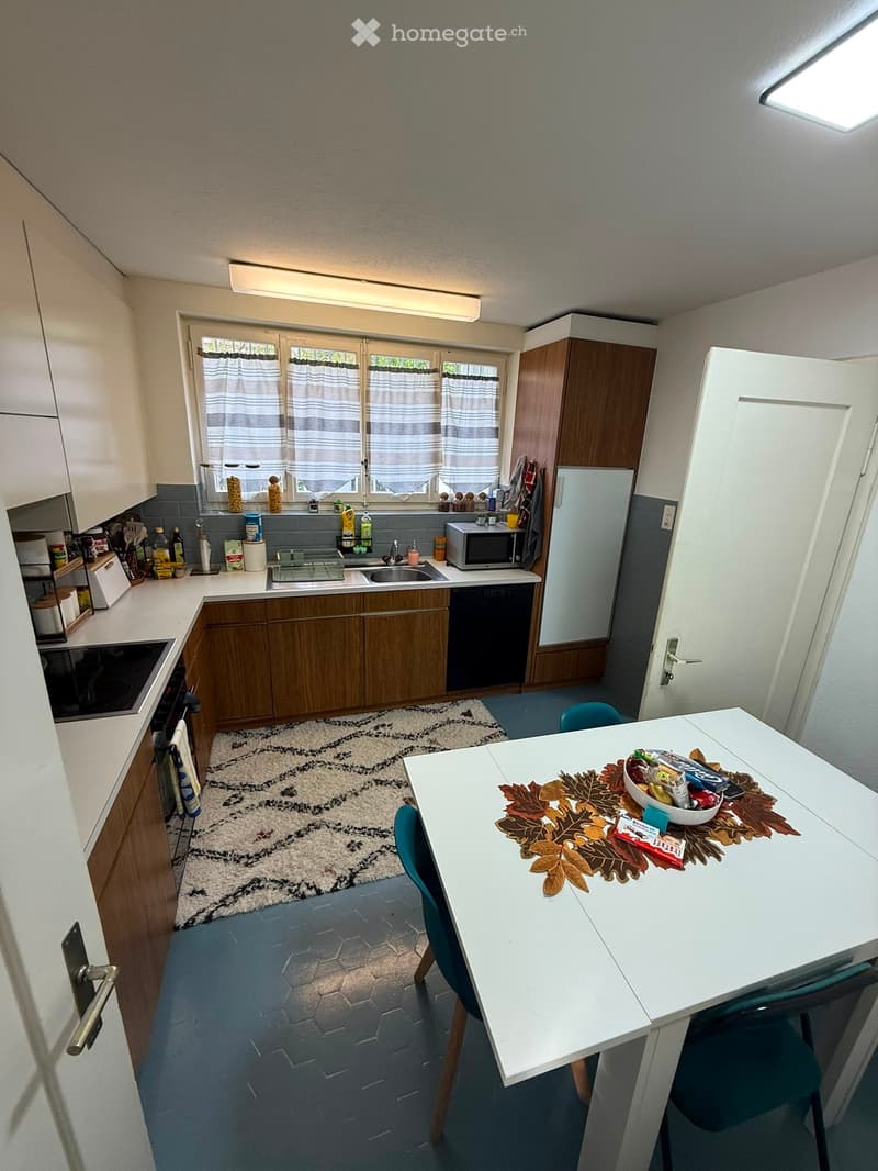 The kitchen features white cabinets, a wooden dining table with chairs, a refrigerator, microwave, sink, and various kitchen items on the counter.
