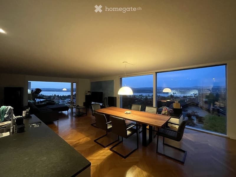 Open-plan living and dining area with large windows offering a scenic view, featuring a sofa, dining table with chairs, and a television.