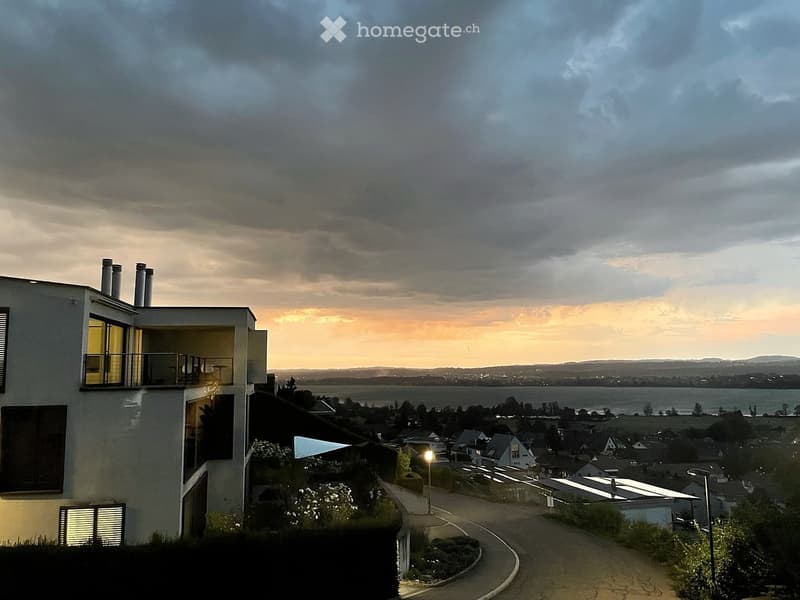 Modern two-story house with large windows and a balcony, overlooking a scenic landscape with a body of water and distant hills.