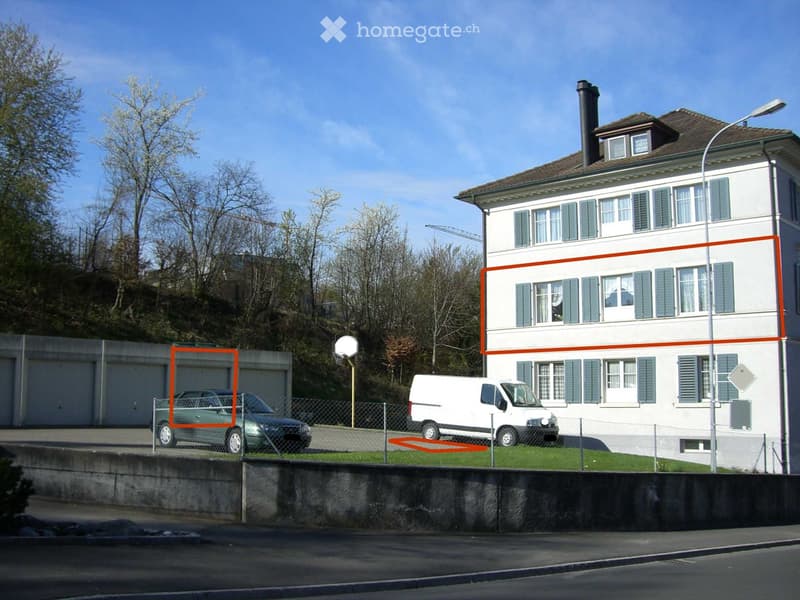 A multi-story residential building with white walls, green shutters, and a brown roof. The building has multiple windows and a chimney. There is a fenced area in front with a parked car, a van, and a basketball hoop.