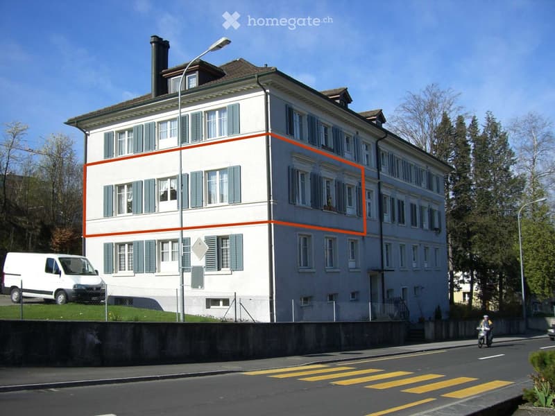 A large, multi-story residential building with white walls, green shutters, and multiple windows. The building has a classic architectural style with a pitched roof and chimney. A white van is parked in front, and the building is situated near a road with