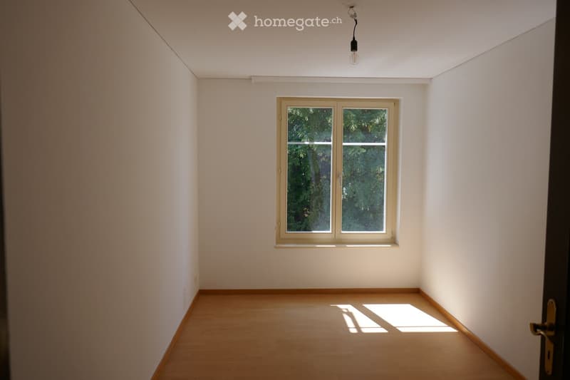 Empty room with white walls, wooden floor, and a single window with a view of greenery outside. Natural light streams in, casting shadows on the floor.