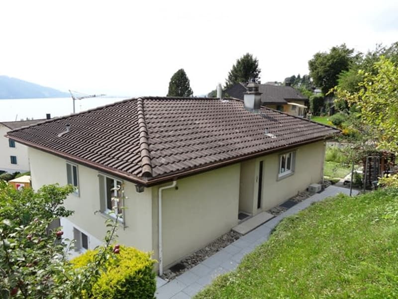 Single-story house with a brown tiled roof, white walls, and a view of a lake in the background. The house has multiple windows and a small garden area with a paved pathway leading to the entrance.