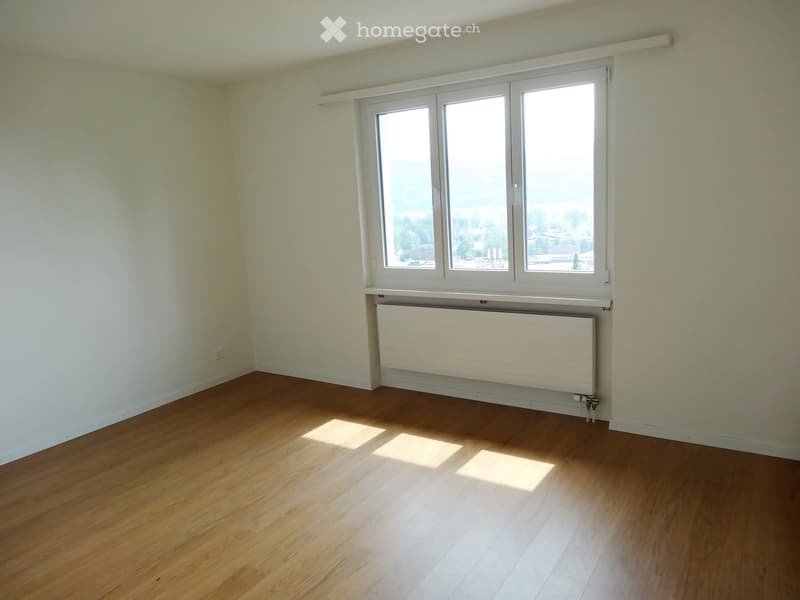 Empty room with wooden flooring, white walls, large window with white frame, and radiator below the window.