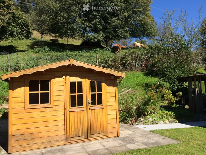 A wooden shed with double doors and windows, situated on a paved area surrounded by greenery and grazing animals in the background.