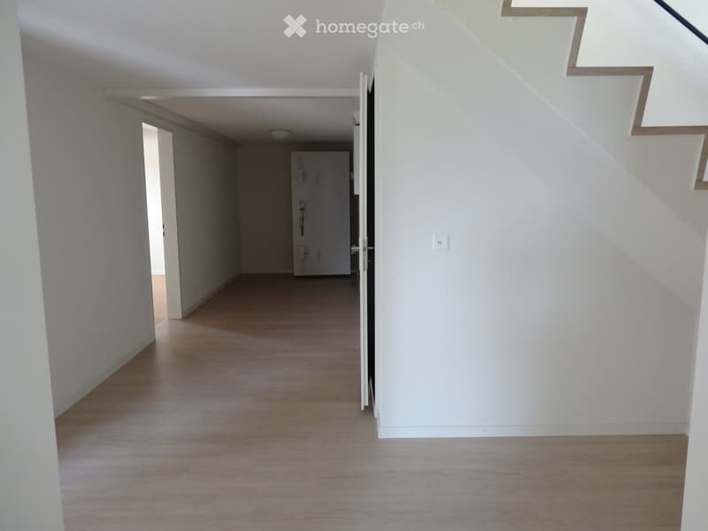 Empty hallway with light wooden flooring, white walls, and a staircase leading upwards. No furniture or decorations are present.