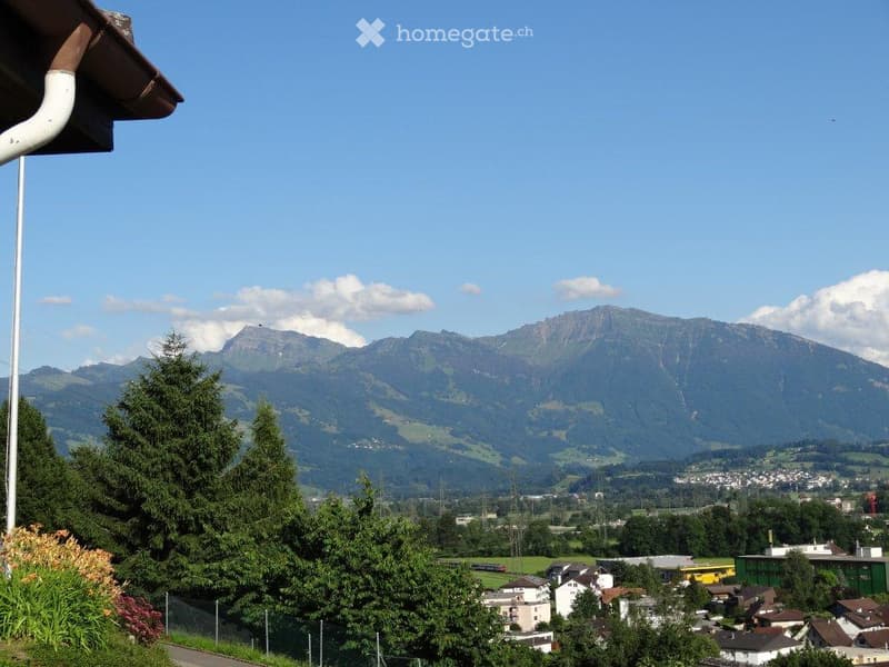 Panoramic view of a town with houses, trees, and distant mountains under a clear blue sky.