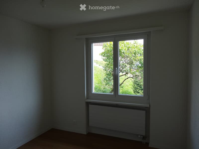 Empty room with wooden flooring, white walls, a large window with a view of greenery, and a radiator below the window.