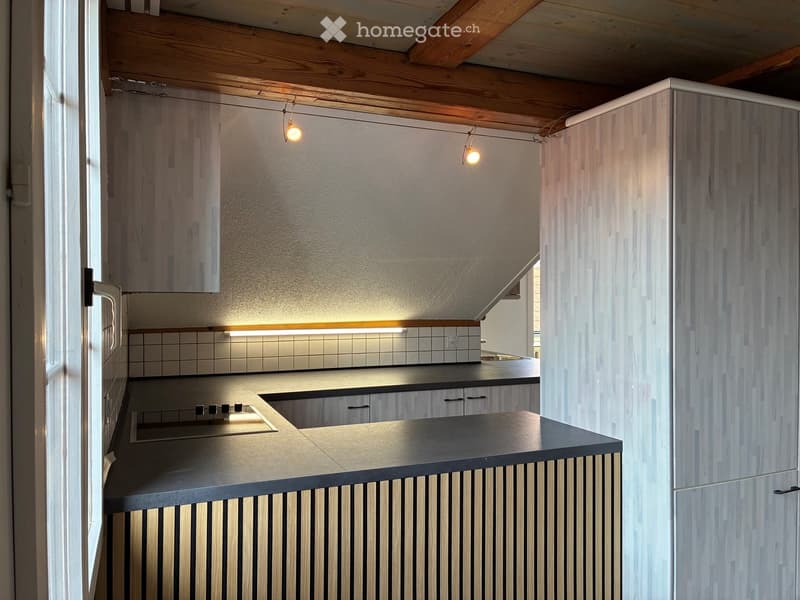 Modern kitchen with sleek wooden cabinets, a black countertop, and built-in appliances. Exposed wooden beams on the ceiling and warm lighting create a cozy atmosphere.