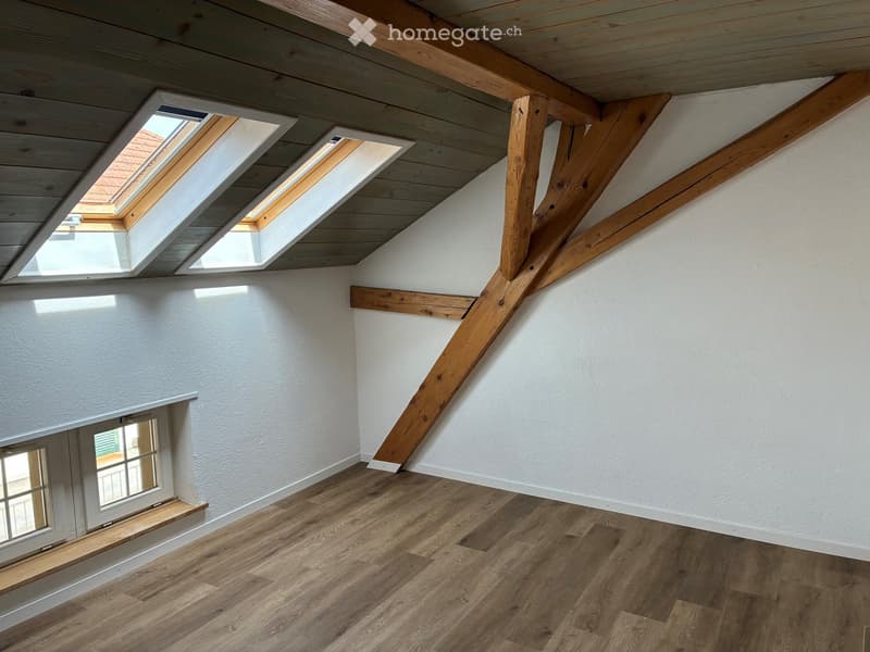 Empty attic space with wooden beams, skylights, and wooden flooring. White walls and exposed wooden beams create a bright, airy atmosphere.