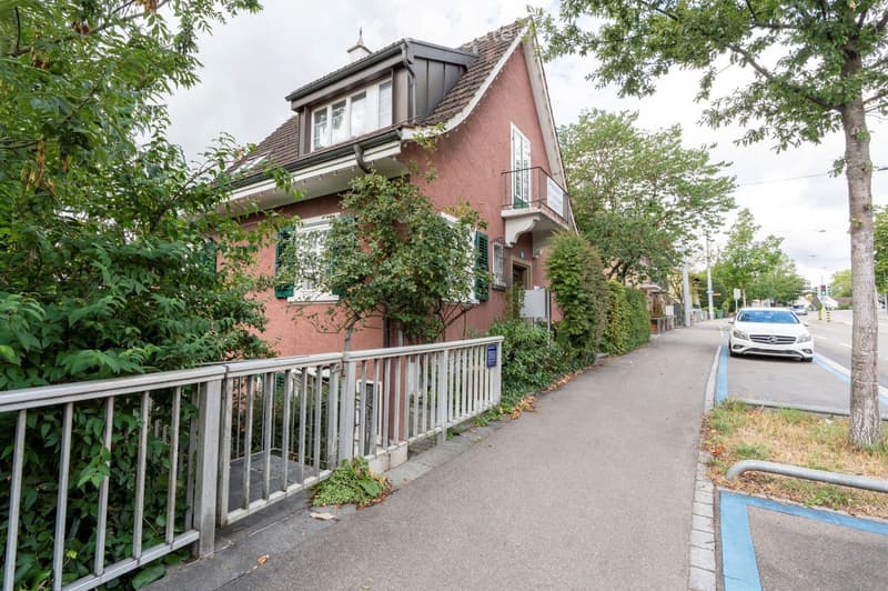 Two-story house with red brick facade, white-framed windows, and a small balcony on the upper floor. The house is surrounded by greenery and a metal fence.