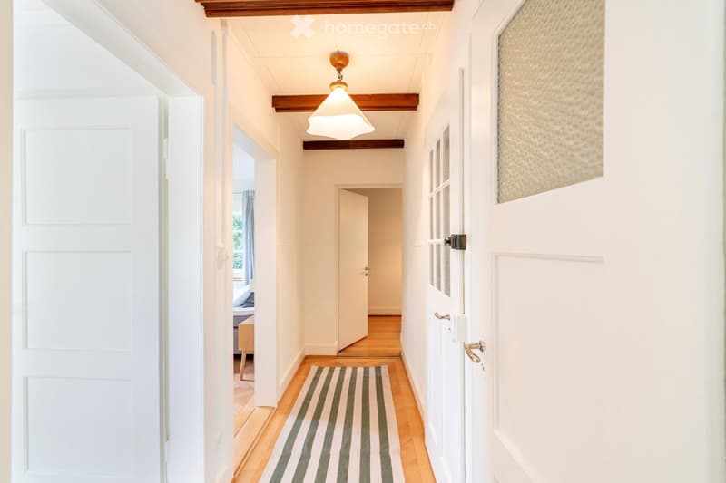 A narrow hallway with wooden flooring, white walls, and a striped runner. Features a hanging light fixture and several doors leading to other rooms.