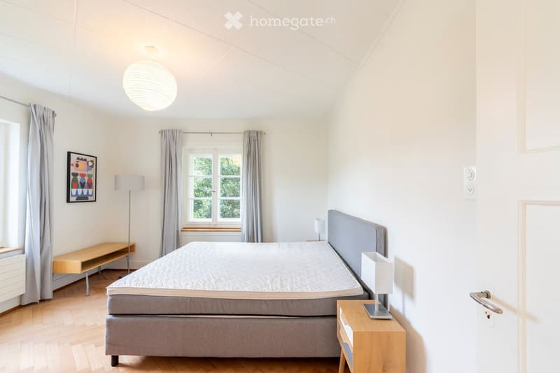 Bright bedroom with wooden flooring, a grey upholstered bed, nightstand with lamp, bench, and a large window with curtains.