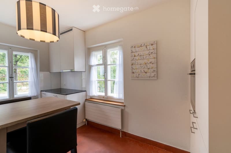 Bright kitchen with white cabinets, a wooden table, and a hanging light fixture. The space features large windows with white curtains and a decorative wall art piece.