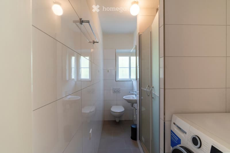 Modern bathroom with white tiles, a toilet, sink, mirror, glass-enclosed shower, and a washing machine in the corner.