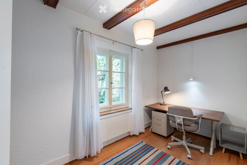 A well-lit study or home office with wooden flooring, white walls, and exposed wooden beams on the ceiling. Features include a large window with white curtains, a wooden desk with a chair, a wall-mounted lamp, and a small side table.