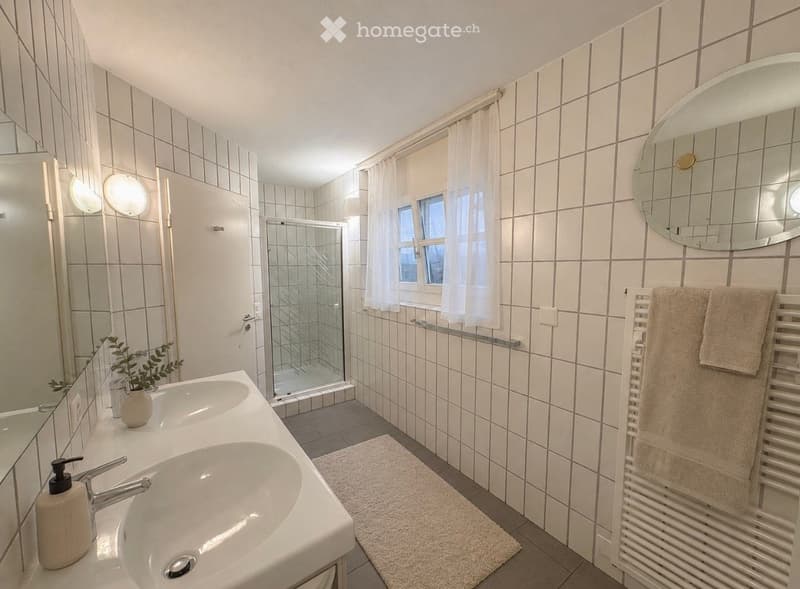 Modern bathroom with white tiled walls, double sink, large mirror, shower area with glass door, and towel rack with towels.