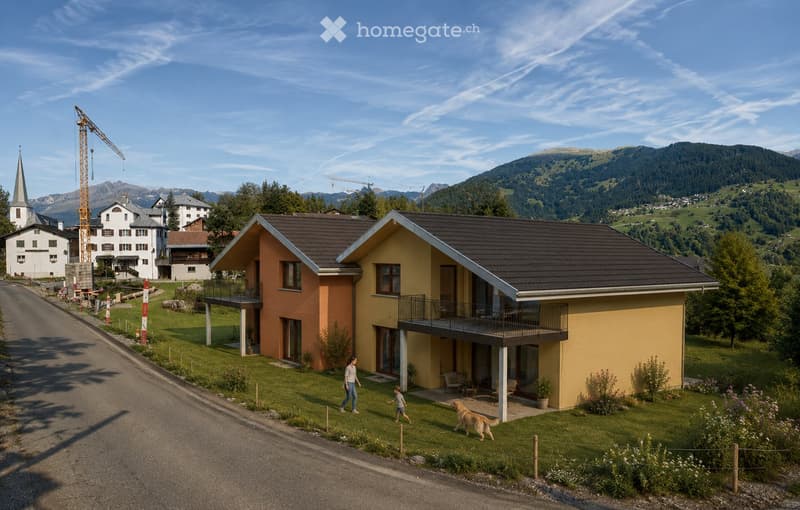 Two-story modern house with yellow and orange exterior, dark roof, and balcony. Located in a scenic valley with mountains in the background.