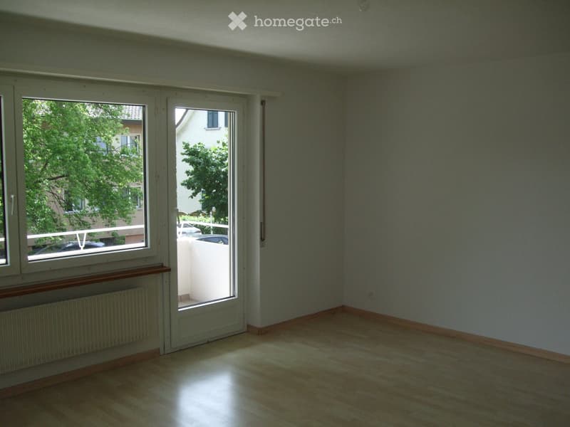 Empty room with wooden flooring, large windows, and a sliding glass door leading to a balcony area.