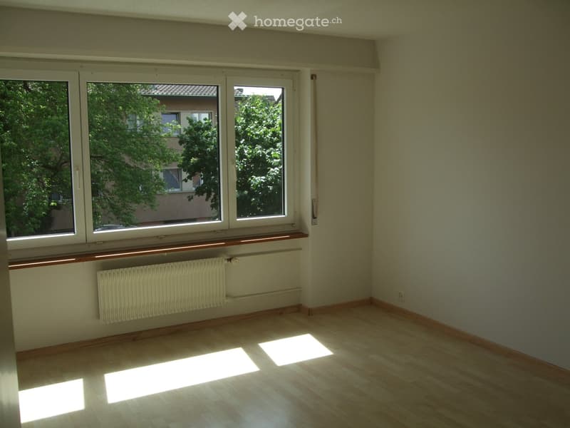 Empty room with wooden flooring, large windows, radiator, and natural light streaming in.