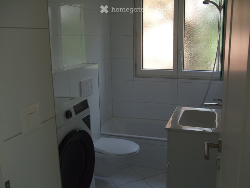 Modern bathroom with white tiled walls and floor, featuring a toilet, sink with faucet, and a washing machine in the corner.