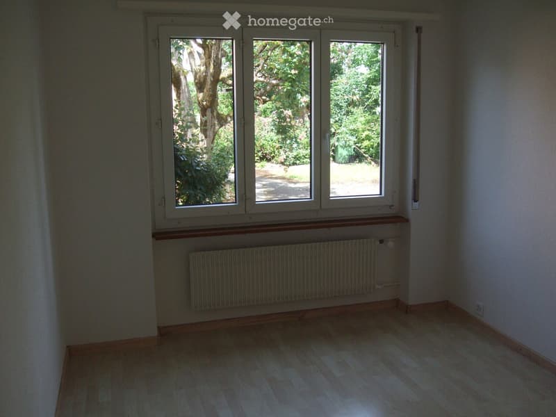 Empty room with wooden flooring, white walls, large windows, radiator, and a wooden shelf below the windows.