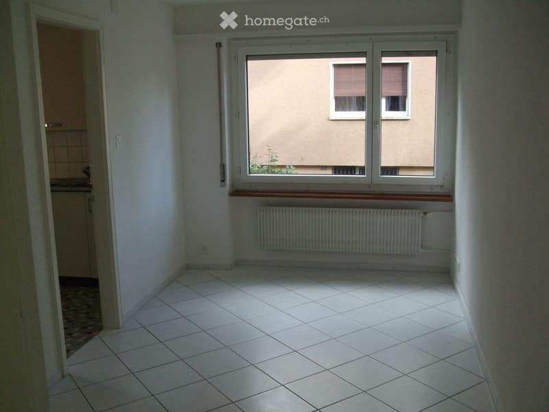 Empty room with white tiled floor, white walls, large window with wooden sill, radiator, and a doorway leading to another room.