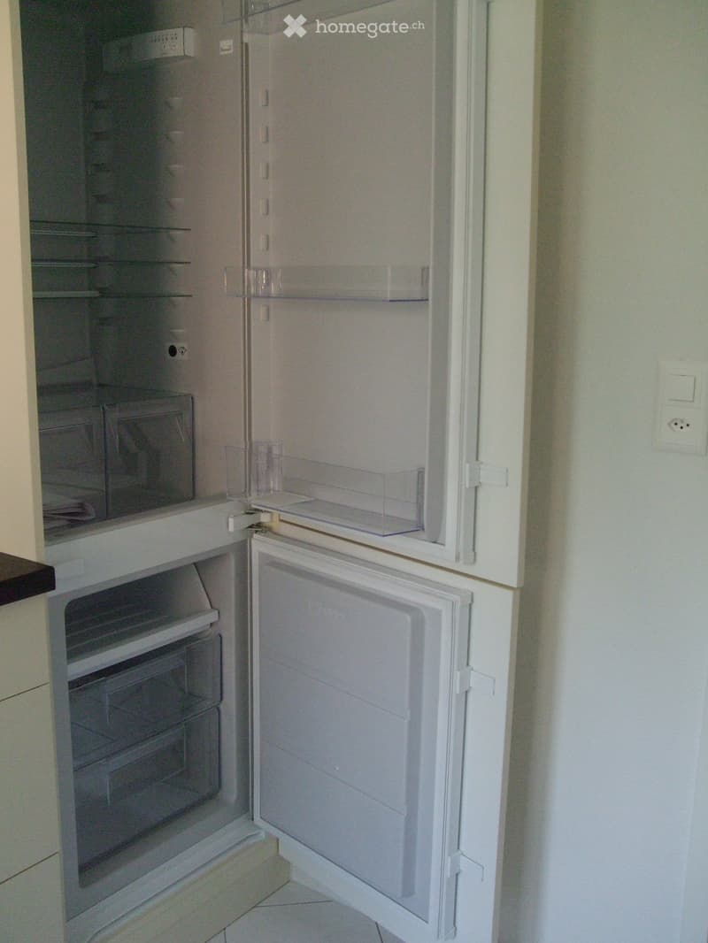 A modern, white refrigerator with multiple compartments and shelves, including a freezer section and a refrigerator section, is shown in a clean, well-lit kitchen area.