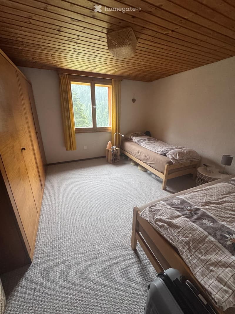 Cozy attic bedroom with wooden beams, two single beds, nightstands, and a large window with curtains overlooking a forested area.