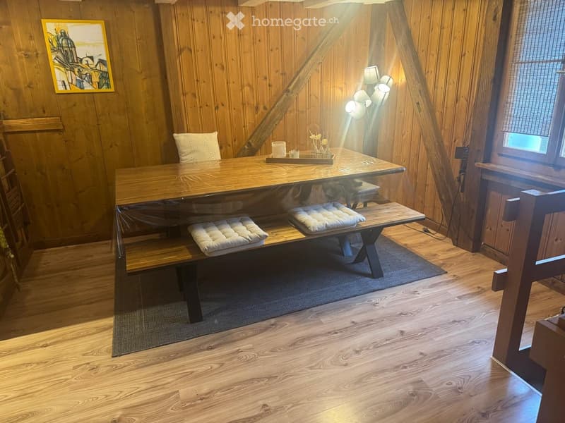 A cozy wooden room with a large wooden table, two benches with cushioned seats, a single chair, a lamp, and a picture frame on the wall.