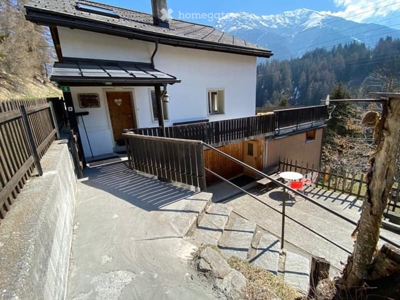A two-story white house with a wooden front door, small balcony, and scenic mountain view in the background.