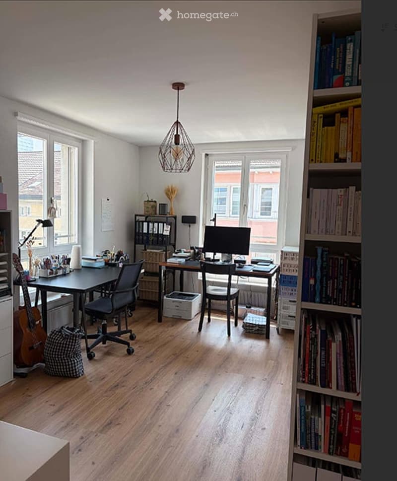 A modern, well-lit home office with wooden flooring, large windows, a desk with a laptop, a comfortable chair, a guitar, and a bookshelf filled with books.