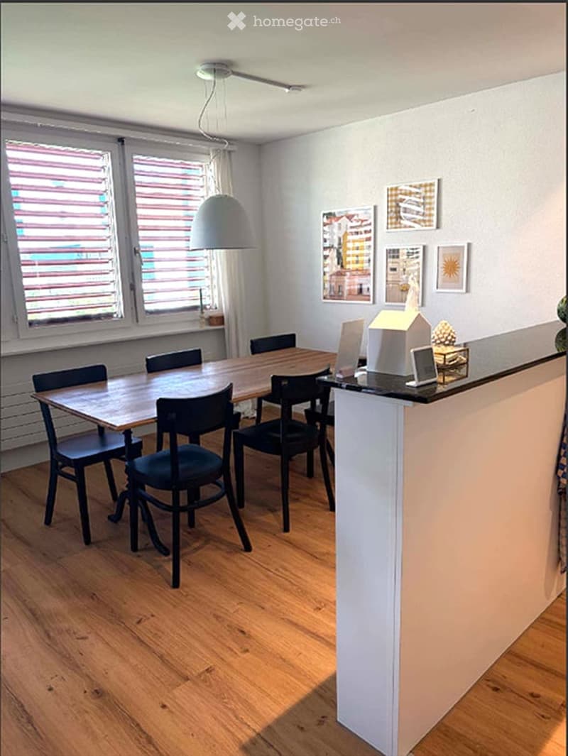 A modern dining area with a wooden dining table, black chairs, a pendant light, and a small kitchen counter with a laptop and decorative items.