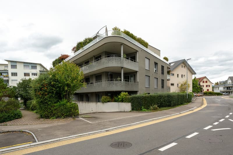 A multi-story building with a green terrace, a balcony with a lacy design, and parking in front.