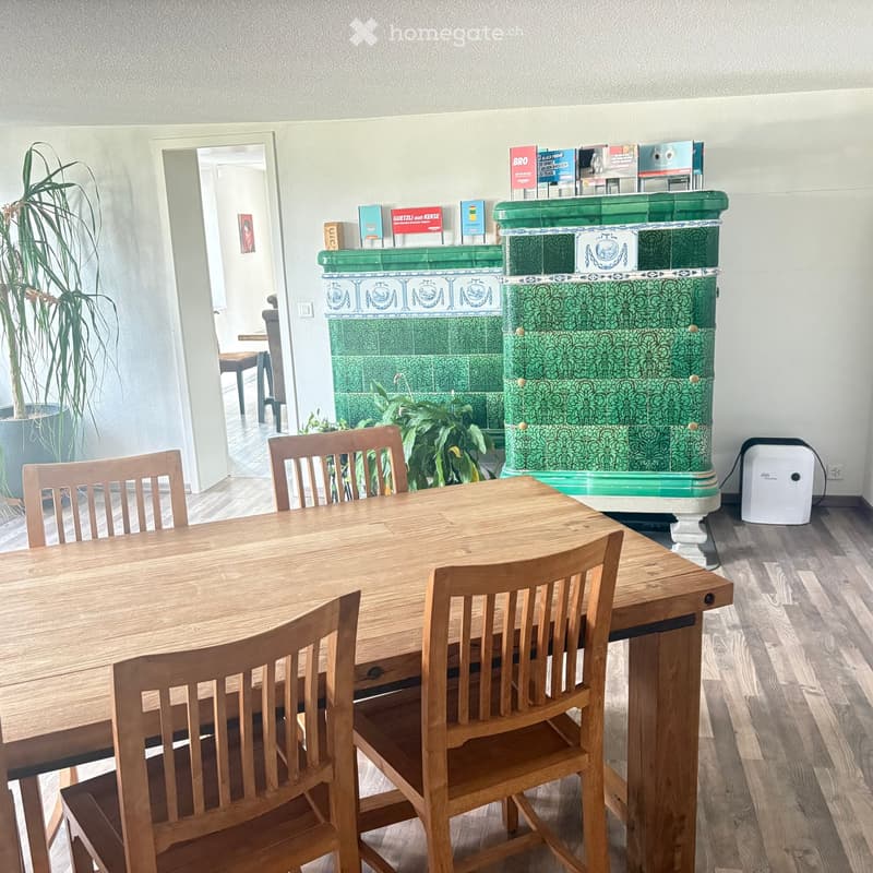 A cozy kitchen with a wooden dining table and chairs, a green-tiled fireplace, and potted plants. The room features light wooden flooring and white walls.
