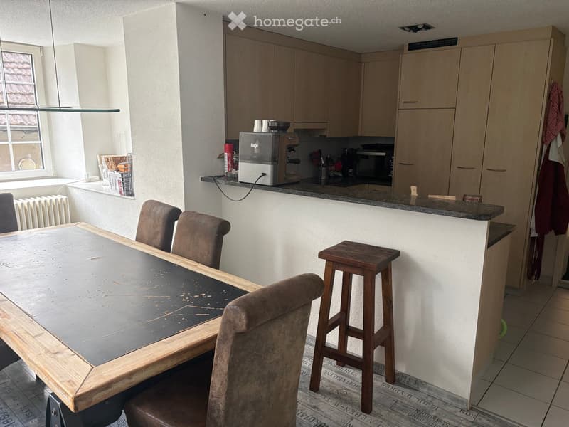 A modern kitchen and dining area with a wooden dining table, chairs, a stool, and kitchen cabinets. A coffee machine and microwave are on the counter.
