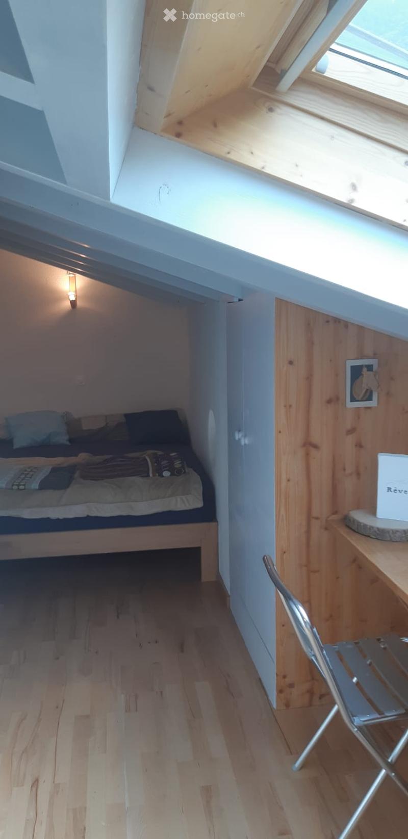 Cozy attic bedroom with wooden floors, slanted ceilings, and a skylight. Features a small bed with pillows and blankets, a wooden table with a chair, and a picture frame on the wall.