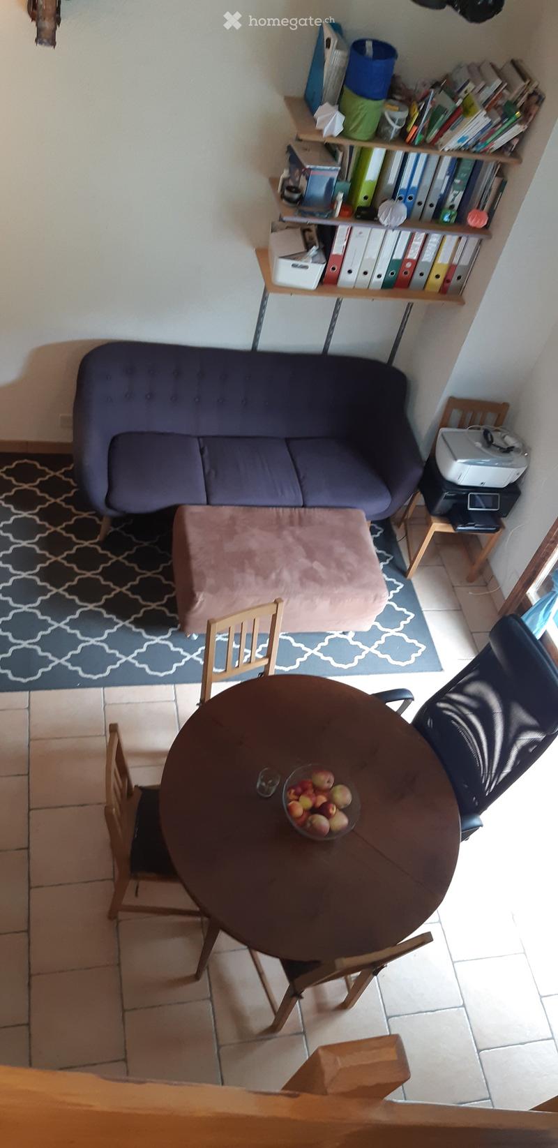 A small, cozy living room with a sofa, a round wooden table with chairs, a bookshelf filled with books and binders, and a printer on a small table. The room features a patterned rug and a cushioned ottoman.