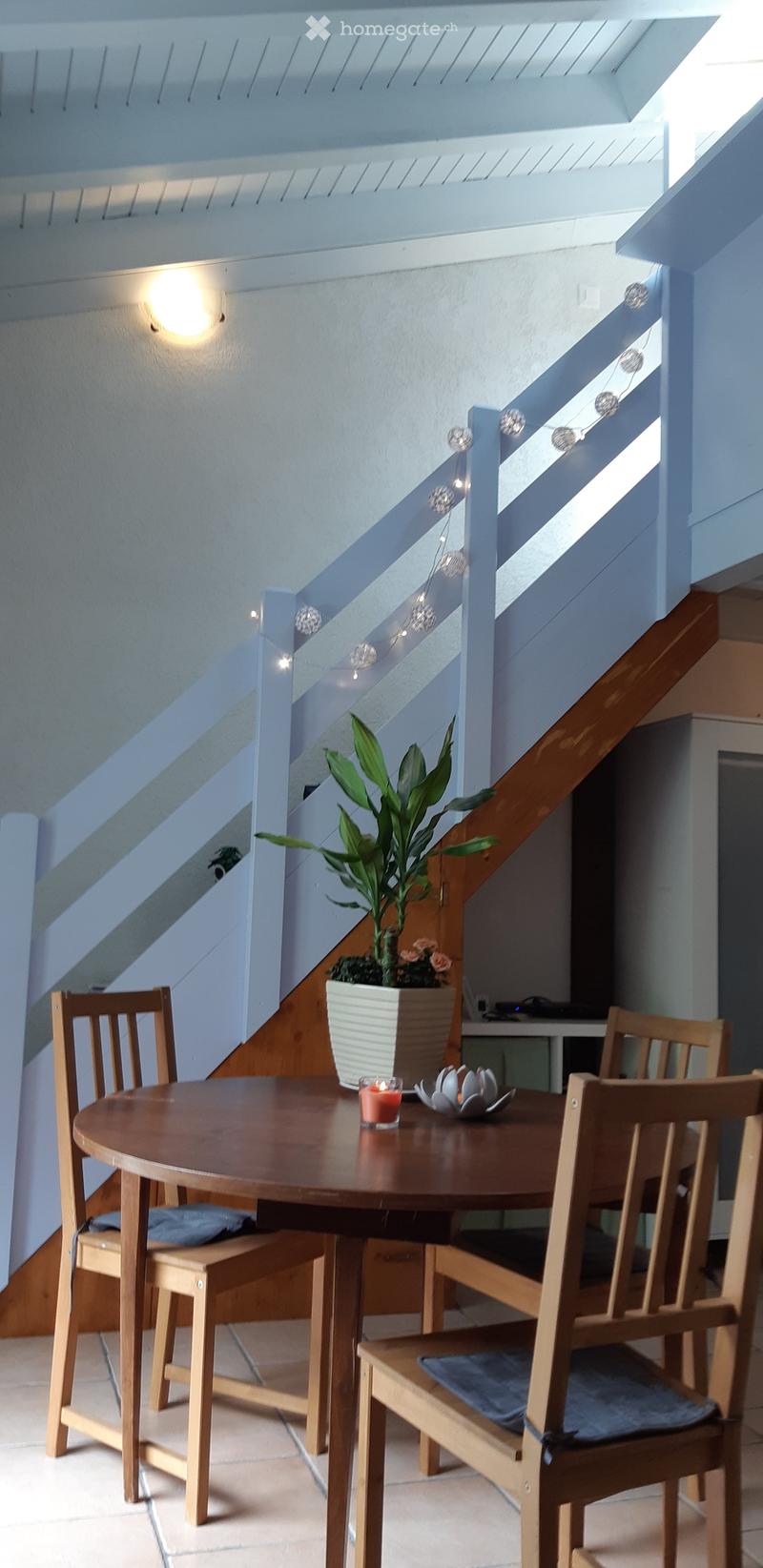 A small, modern dining area with a round wooden table, four wooden chairs, a potted plant, and a lit candle on the table. The area is adjacent to a staircase with a glass railing and string lights.