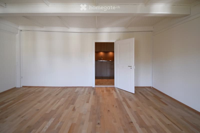 Empty room with wooden flooring and white walls, featuring an open doorway leading to a small kitchen area.