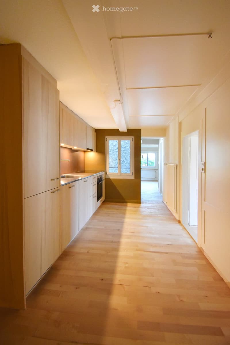Bright hallway with wooden flooring, white walls, and a small kitchen area visible on the left side. Natural light streams in from a window at the end of the hallway.
