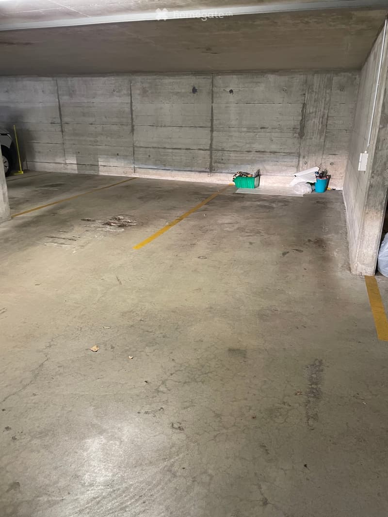 Empty underground parking space with concrete walls and floor, marked parking lines, and a few scattered items in the corner.