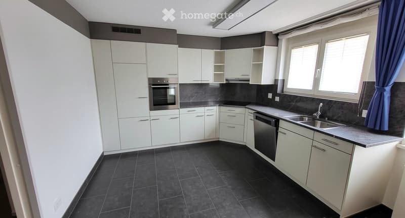 Modern kitchen with white cabinets, dark countertops, and stainless steel appliances including an oven and dishwasher. Features a large window with blue curtains and dark tiled flooring.