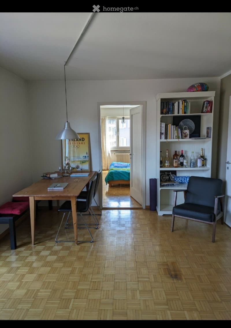 A well-lit room with wooden flooring, a dining table with chairs, a bookcase, a mirror, and a lamp. An adjacent bedroom is visible through an open doorway.